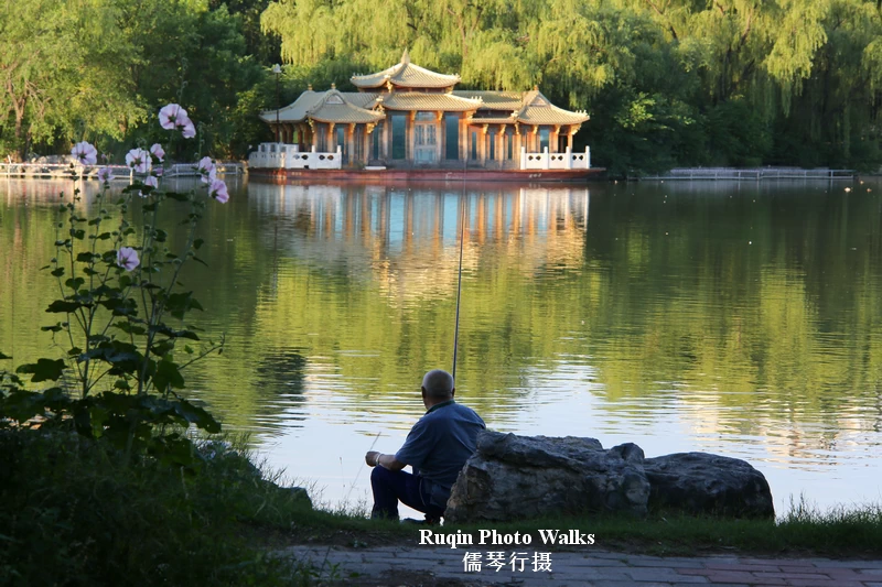 A white-haired man fishing quietly by the lakeshore, with traditional buildings reflected across the water in soft morning light and greenery in Longtan Lake Park
